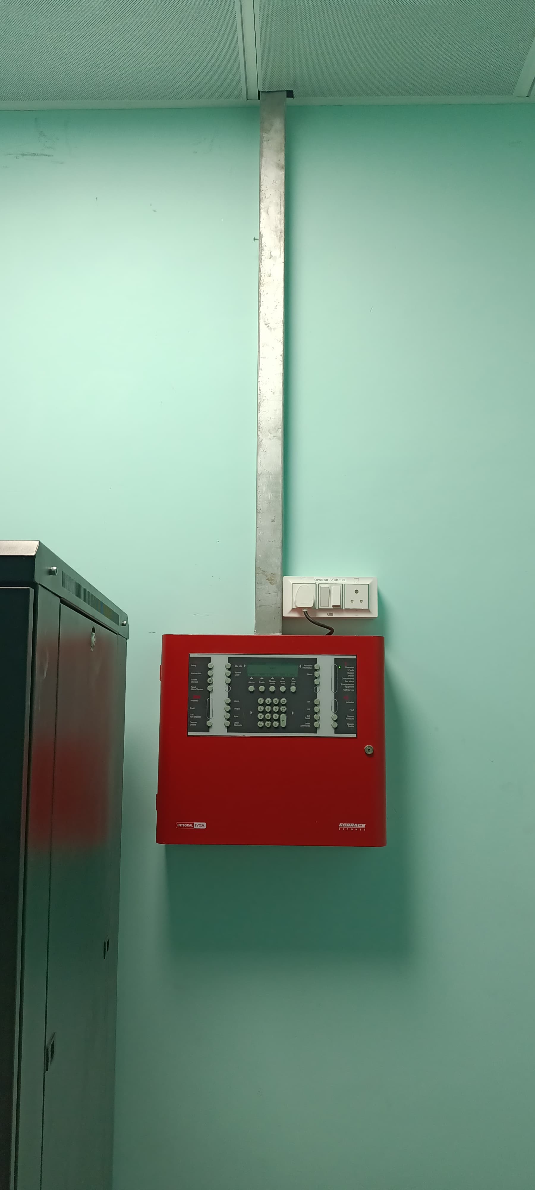 Red Schrack Seconet fire alarm control panel on a light green wall with metal conduit.